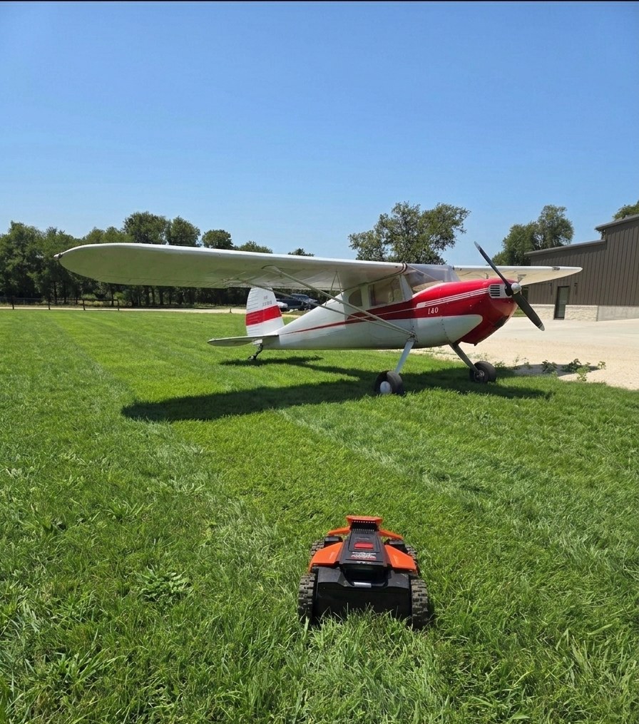 Tracked robotic mower cutting a grass airstrip beside a red and white Cessna 140 taildragger
