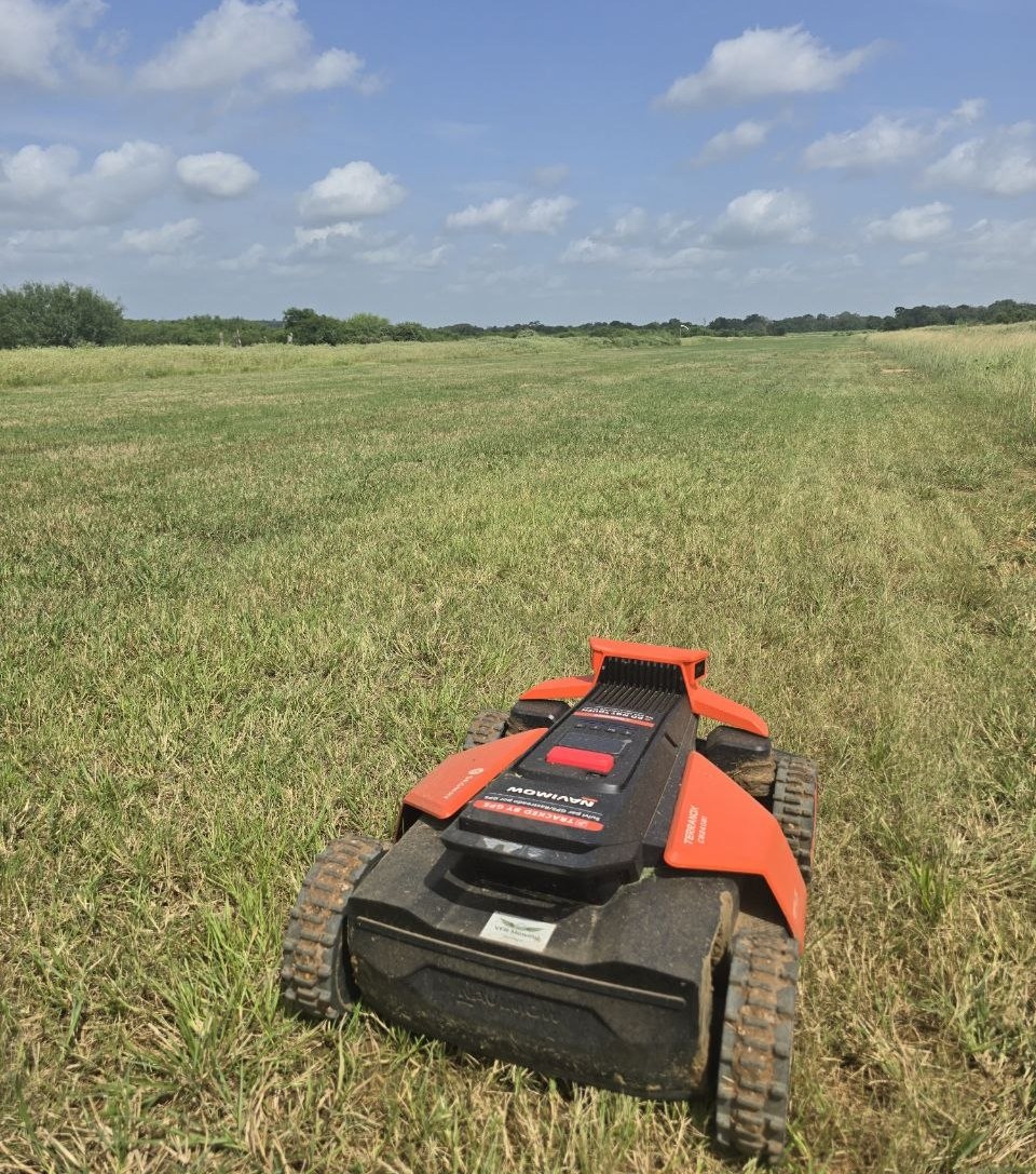 Orange tracked robotic mower cutting a wide grass runway under a blue Texas sky