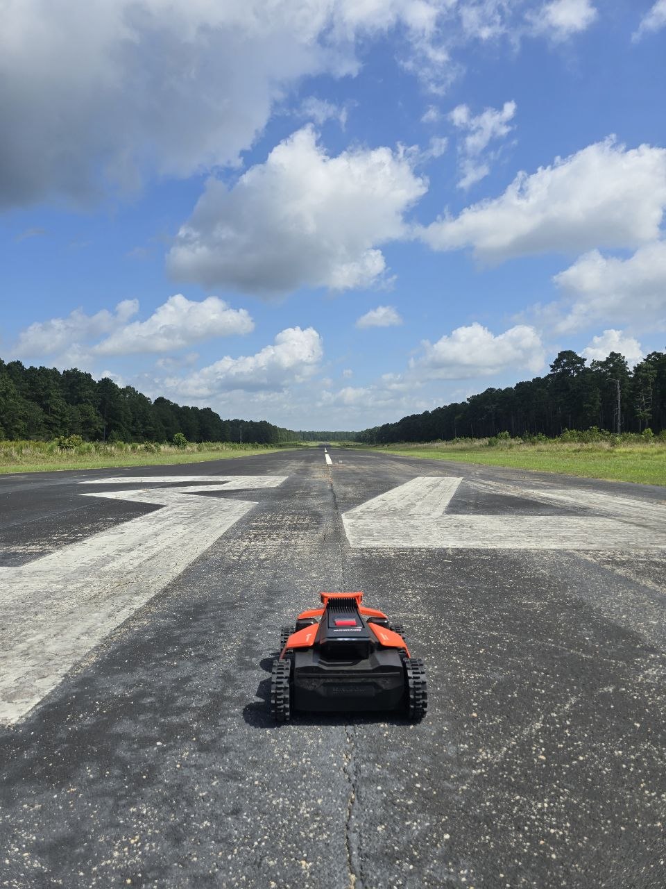 Orange tracked robotic mower parked on a paved runway with the 34 threshold markings, the runway stretching to the horizon under a blue sky